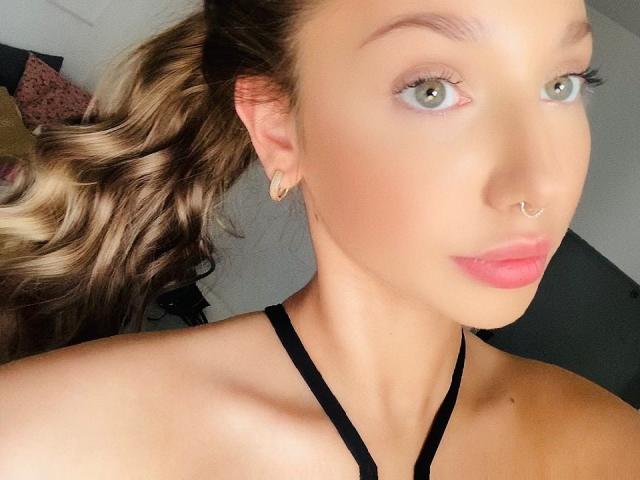 EmmaPassionate with light green eyes, pink lips, and a nose ring, wearing a black halter top, has wavy blonde hair in a ponytail, and small gold earrings.
