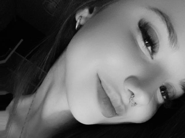 EmmaPassionate's face in a black-and-white selfie, showing her nose ring, subtle smile, and long eyelashes. Her ear has a small stud, and her hair is partially visible.