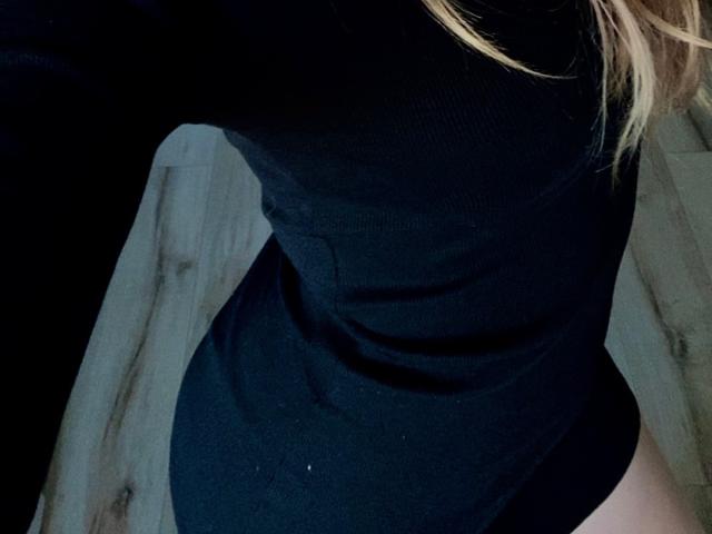 EmmaPassionate in a black, form-fitting, long-sleeve top, with blonde hair visible, showing her small, round ass and a hint of her bare, pale, upper thigh.