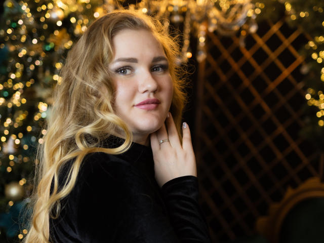 MikeAndAnna with long blonde curly hair, black velvet top, looking at camera, slight smile, hand near face, bokeh lights in background, soft glow, gentle expression, cute face.