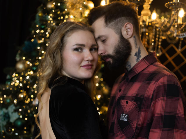 MikeAndAnna with wavy blonde hair, black velvet dress, and MikeAndAnna with dark beard, plaid shirt, and tattoo, nose-to-nose, looking at camera. Christmas tree with lights in background.