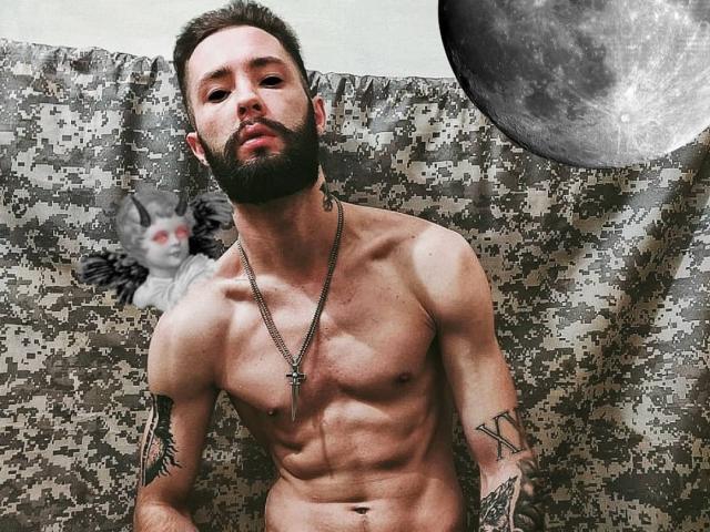 MikeAndAnna is a shirtless, bearded, tattooed dude with a silver cross necklace, muscular, and looking down, standing in front of a pixelated pattern with a gray stuffed cat.
