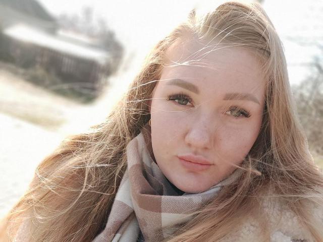 MikeAndAnna has light brown hair, green eyes, and a small nose with a slight pout. She's wearing a plaid scarf and a fluffy coat, with wind in her hair.