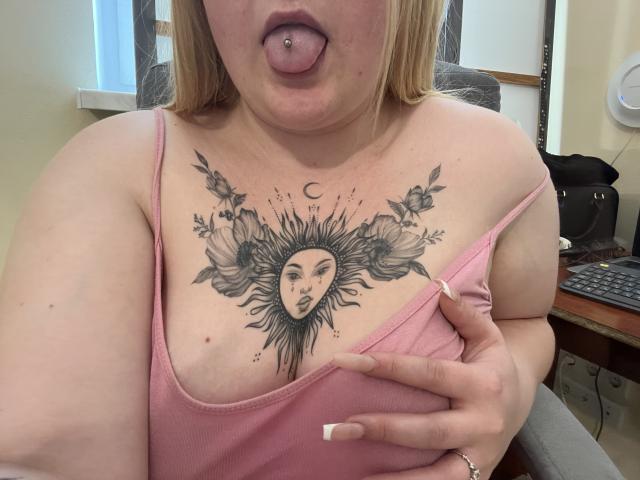 DiLaria with blonde hair, tongue out, pink tank top, holding her tits, sun/moon tattoo on chest, nipple piercing, pink nails, sitting in office chair, desk in background.