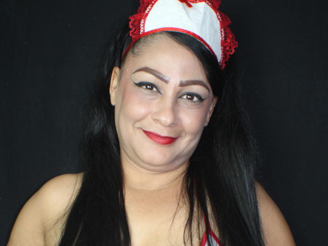 CandelaFoxy, con cabello negro largo, lleva un gorro de enfermera rojo y blanco, sonriendo a la cámara, mostrando su lápiz labial rojo y delineador de ojos. Tiene un rostro curvilíneo y regordete..