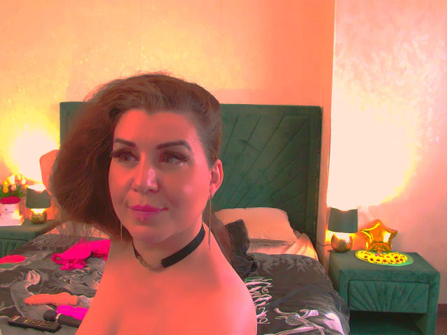 RosseFiftyThree with pink lipstick, dark green choker, and shoulder-length brown hair, sitting topless on a bed with a green headboard, pink bra, and star-shaped ornament.