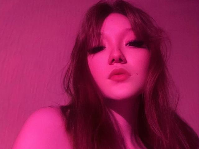 AmberAura's face, pink-lit, with long brown hair, heavy eyelashes, and slightly parted lips, showing a small beauty mark on her right cheek. Her nose is pierced.