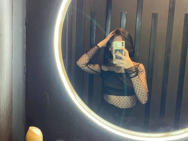AmberAura with hands in brown hair, wearing see-through black polka-dot top, mirror selfie, showing tits, holding phone, round mirror with neon ring, yellow bottle bottom left.