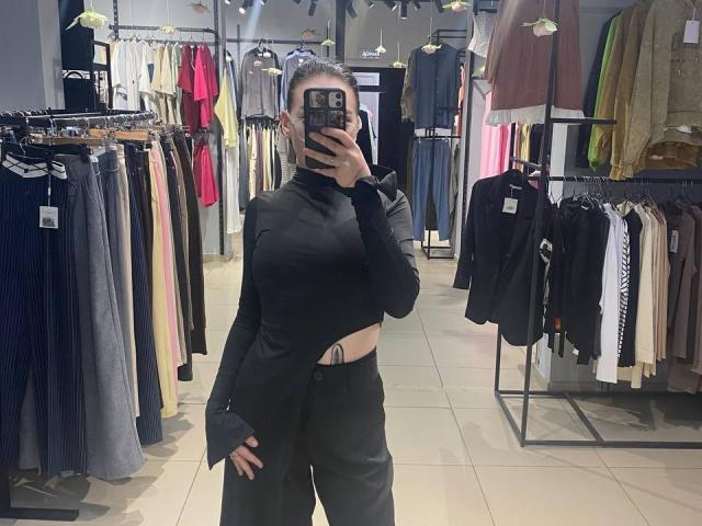 AmberAura, black long-sleeve top, high-waisted black pants, visible tattoo on right side, taking mirror selfie in clothing store, holding phone with black case, blonde hair in bun.