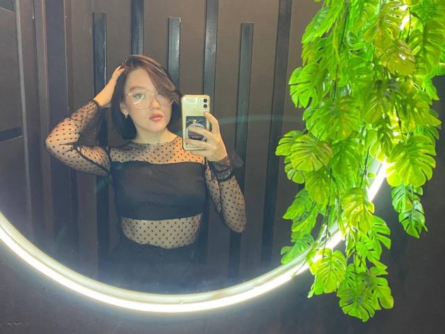 AmberAura with glasses, holding phone, wearing sheer black polka dot top, black bralette, dark hair, hand in hair, mirror selfie, green leafy plant, ring light, mirror reflection.