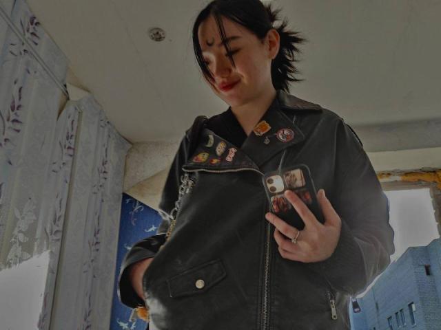 AmberAura with dark hair in a messy ponytail, wearing a black leather jacket with patches, holding a phone, smiling slightly, standing in a dimly lit room.
