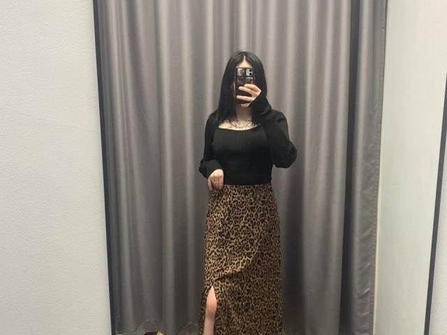 AmberAura with black top, leopard skirt, thigh slit, holding phone, mirror selfie, black straight hair, tattoos on chest, gray curtain, standing, white wall, taking photo.