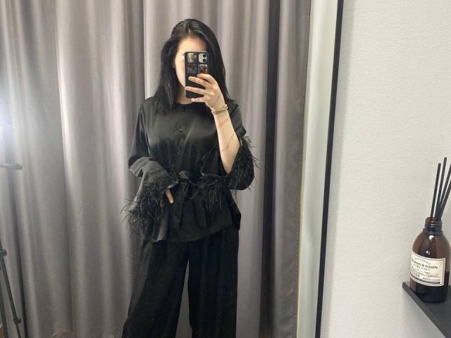 AmberAura with black frilled shirt, black pants, holding phone in mirror selfie, long black hair, white wrist, standing, gray curtain background, black shelf with brown bottle.