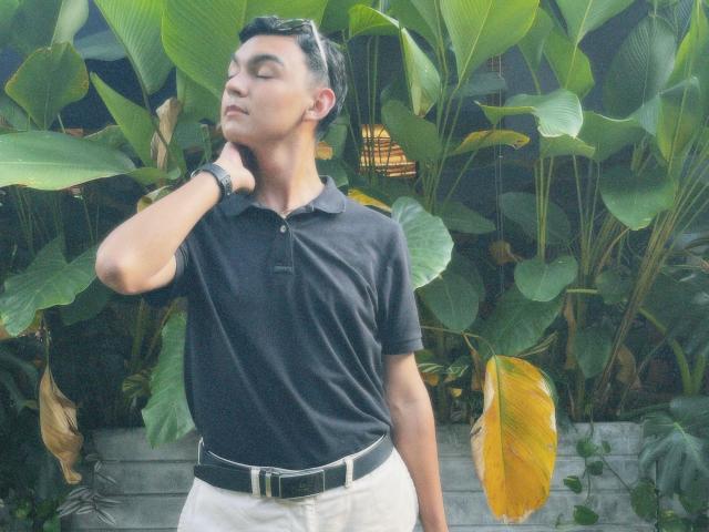 DeusBeauty with short black hair, wearing a black polo and white pants, has a black belt, wristwatch, and is touching his neck with eyes closed, standing in front of green plants.
