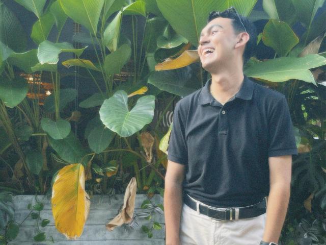 DeusBeauty, a cute Asian dude with short black hair, wearing a black polo and white pants, smiling up at green leafy plants, with sunglasses on his head.