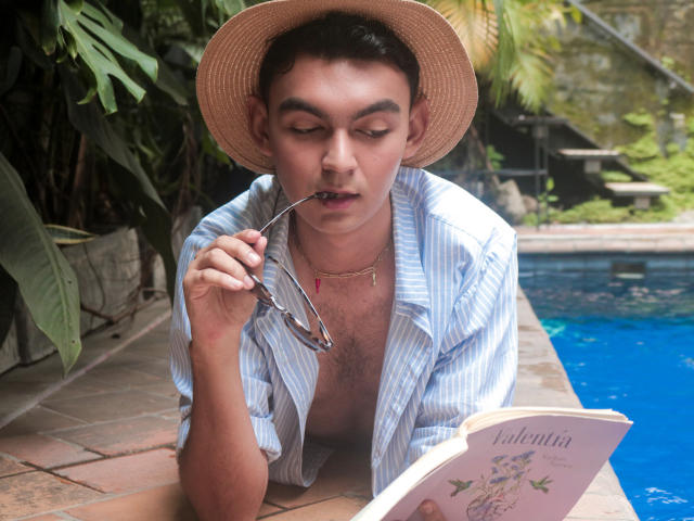 DeusBeauty, wearing a straw hat, blue striped shirt, and no pants, leans by a pool, holding glasses in mouth, reading "Valentina" book, chest hair visible, brown eyes.