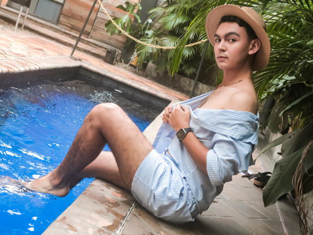 DeusBeauty with tan skin, short dark hair, and a hat, sitting by a blue pool, wearing an off-shoulder striped shirt, exposing a shoulder, and a smartwatch.