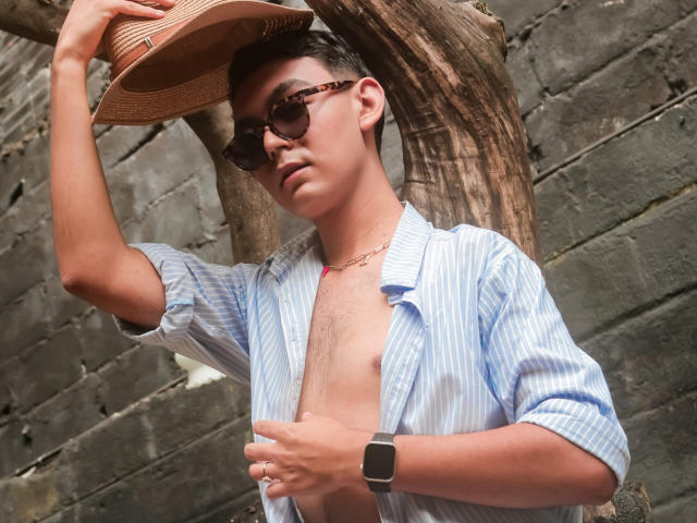 DeusBeauty, shirt unbuttoned, chest visible, wearing a straw hat, dark sunglasses, and a smartwatch, holding hat with left hand, standing by a tree, wearing a necklace.