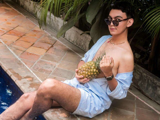 DeusBeauty with dark curly hair, glasses, and a watch, wearing a loose blue shirt, holding a pineapple by the pool, sitting on stone tiles with legs in water.
