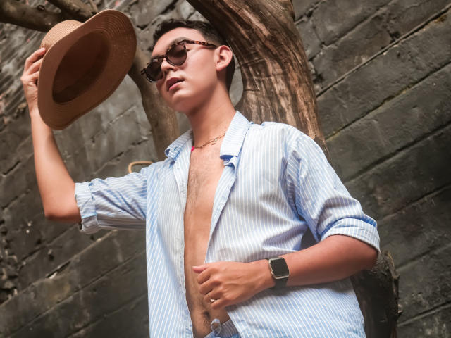DeusBeauty wears a blue-striped shirt, open chest, holding a straw hat, wearing sunglasses, and a watch, standing by a tree. Packed with a backpack, looking cool.