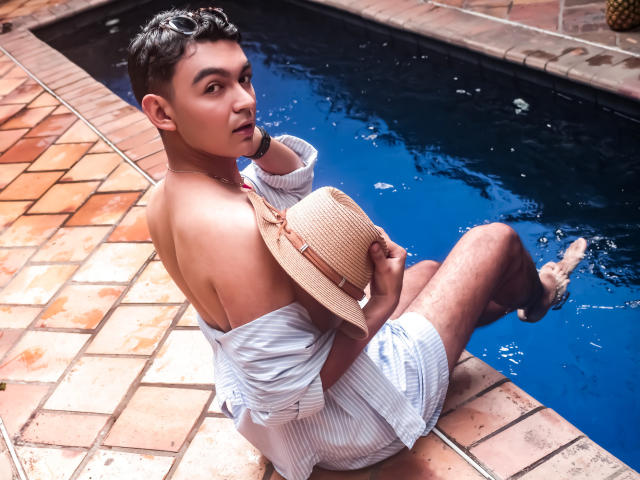 DeusBeauty, shirt off, white striped shorts, sitting by blue pool, holding straw hat, looking back, short dark hair, tan skin, muscular, sexy, tiled poolside, relaxed, summer vibe.