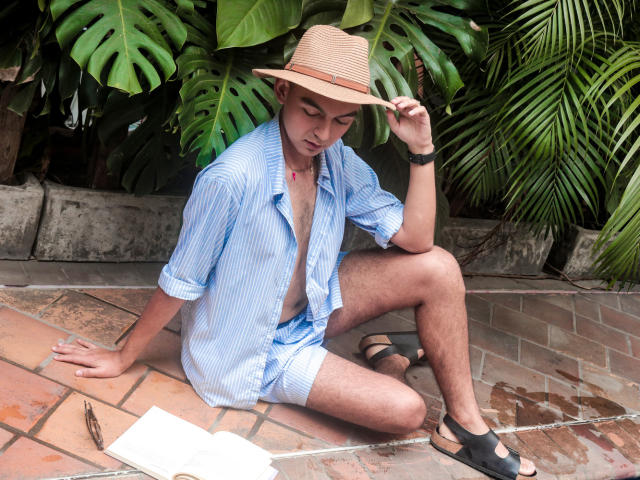 DeusBeauty, a dude with a tan, wearing a blue striped shirt, no pants, black sandals, and a straw hat, sitting on a tiled patio, holding the hat.