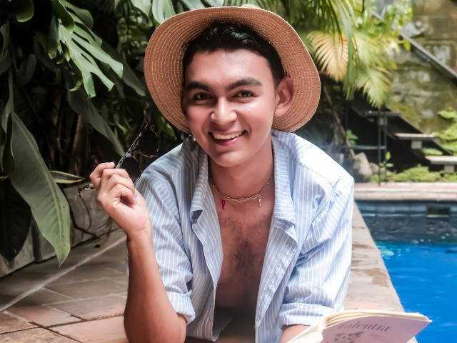 DeusBeauty with short dark hair, tan skin, smiling, wearing a straw hat, blue striped shirt, holding sunglasses, by a pool, chest hair visible, book in hand.