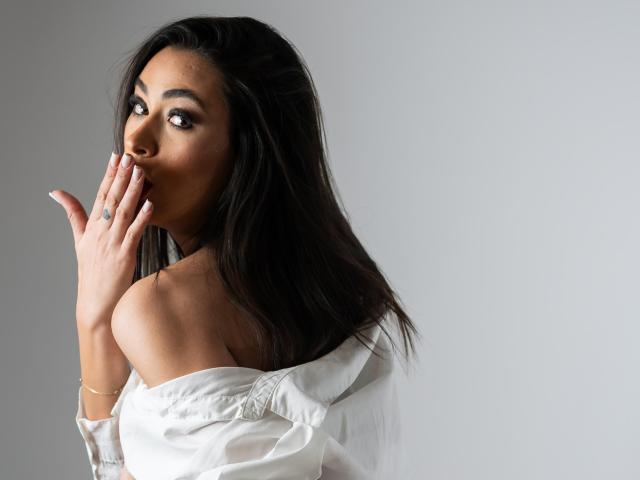 PrettyLiaa with dark, wavy hair, brown skin, and a cute tattoo on her finger, wearing a white off-shoulder shirt, touching her lips with a shy, sexy expression.