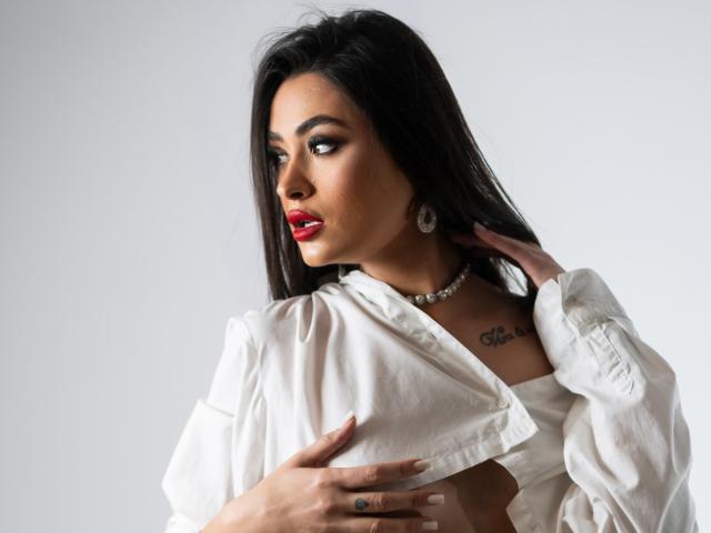 PrettyLiaa with long black hair, red lipstick, white shirt, necklace, and tattoo, holding shirt open, showing chest, looking to side, hand on neck, ring on finger.