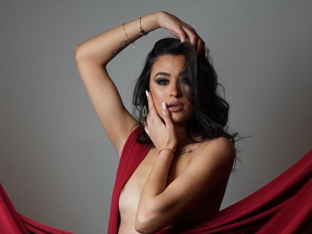 PrettyLiaa with dark wavy hair, wearing a red dress, exposing her tits, arms raised, hands touching her face, gold bracelet, tattoo on chest, grey background, seductive gaze.
