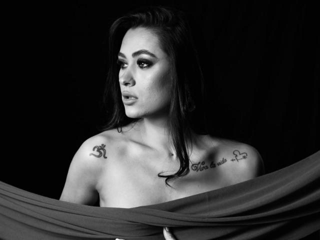 PrettyLiaa with tattoos on her shoulders, topless, dark hair, serious expression, draped in a gray fabric, black and white photo, visible collarbone tattoo, looking to the side.