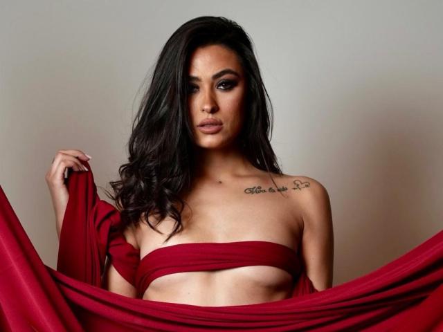 PrettyLiaa with long dark hair, holding a red cloth over her tits, exposing her small ass and a tattoo on her right shoulder. She looks seductive and confident.