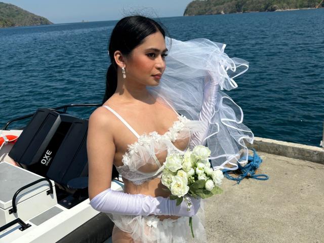 GodessSahara with black hair, wearing sheer white lingerie, floral bust, white gloves, and veil, holding white roses, on a boat, with a bag and blue rope in background.