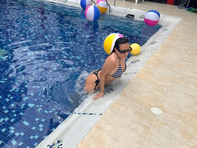 Darablowertg in a blue-striped bikini, holding a yellow and pink beach ball, kneels in a swimming pool with water splashing around her butt, wearing dark sunglasses.