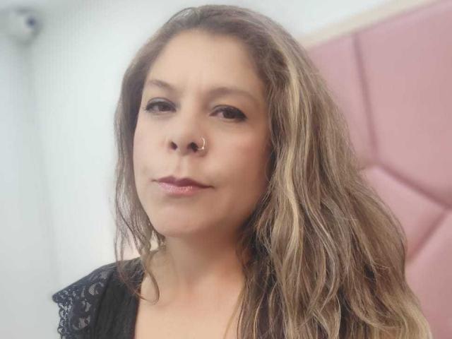 MartinaFiore with wavy brown hair, nose ring, and glossy pink lips, wearing a black lace top, looking straight into the camera with a subtle, confident smile.