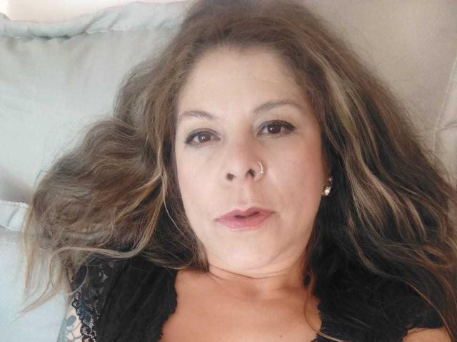 MartinaFiore's face, with brown wavy hair, nose ring, and black lace top, lying on a pillow, showing a neutral expression and light makeup.