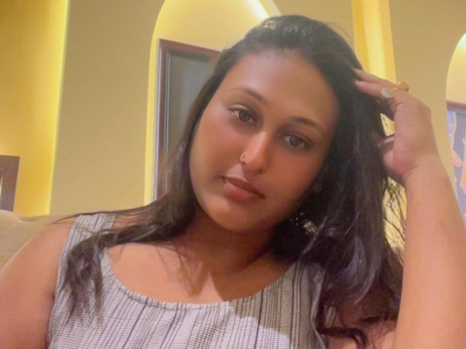 HotLoverForYou with long dark hair, tan skin, brown eyes, wearing a checkered top, hand in hair, gold ring, slight smile, looking at camera, in a yellow-lit room.