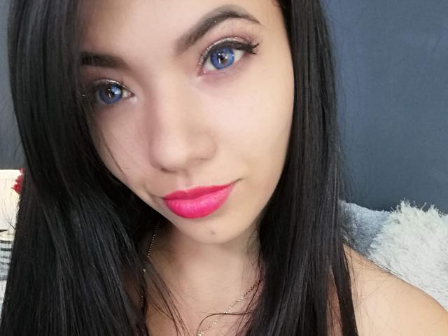 SofieWhite has bright blue eyes, long black hair, and pink lipstick. Her face is slightly shiny, with a small mole below her nose. She's wearing a thin gold necklace.