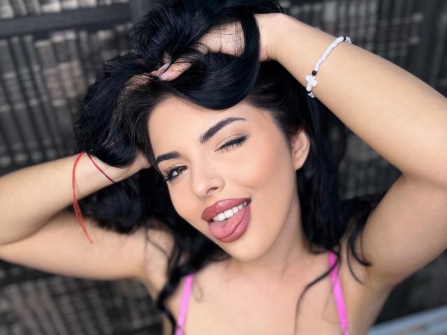 CelineKimberly with black hair, hands in hair, tongue out, pink bra, white bracelet, red thread, smiling, eyebrows, makeup, and medium-sized tits in a photograph.