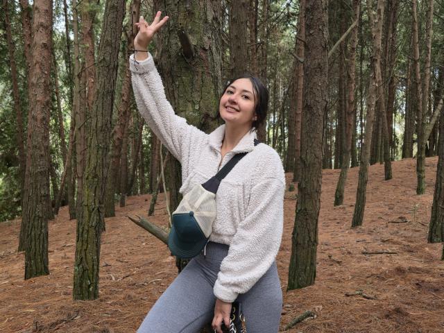 AmberxOne, con i capelli scuri, una giacca bianca e pelosa, pantaloni grigi e una borsa verde, alza la mano destra, sorridendo in una foresta di pini, mostrando un segno di pace con le dita..