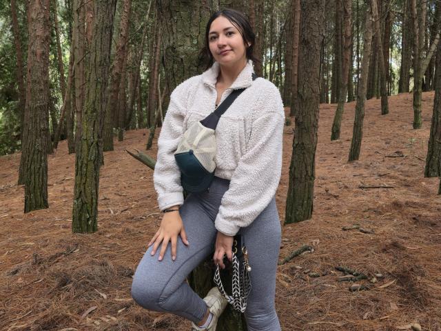 AmberxOne tem uma jaqueta branca e fofa, calças cinzas e uma bolsa transversal, e está em uma floresta com pinheiros. Ela está sorrindo, com uma mão no joelho..