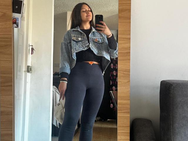 AmberxOne, con lunghi capelli castani, indossa una giacca di jeans, una maglietta nera e pantaloni attillati grigio scuro, è in piedi davanti a uno specchio, tiene in mano un telefono e mostra il suo sedere..