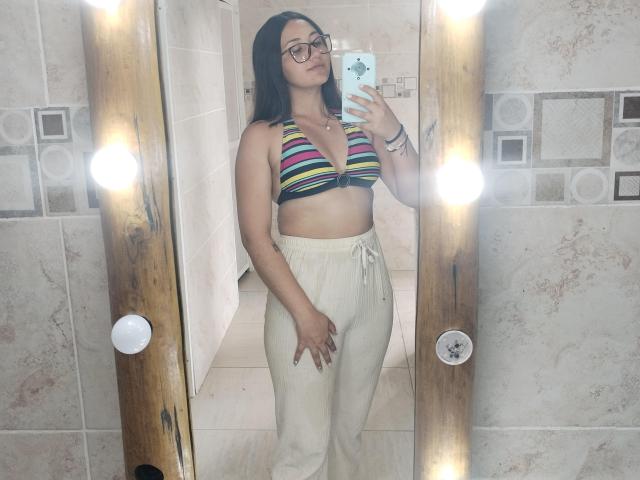 AmberxOne avec des lunettes, haut de maillot de bain rayé laissant entrevoir ses seins, pantalon blanc, tenant un téléphone, selfie devant un miroir, miroir en bois avec des lumières, peau claire, cheveux noirs, tatouage au poignet droit..