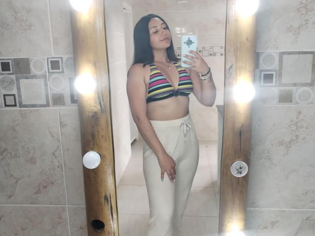 AmberxOne tiene el pelo oscuro, lleva un top de tirantes a rayas que deja ver sus pechos y pantalones de color crema, está tomando una foto de sí misma frente a un espejo con un teléfono, sonriendo, en un baño con azulejos..