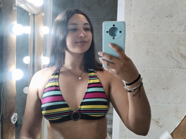 AmberxOne, avec des cheveux bruns, portant un haut de maillot de bain à rayures colorées, tenant un téléphone, souriant, montrant ses seins, et portant des bracelets, dans un selfie pris devant un miroir..