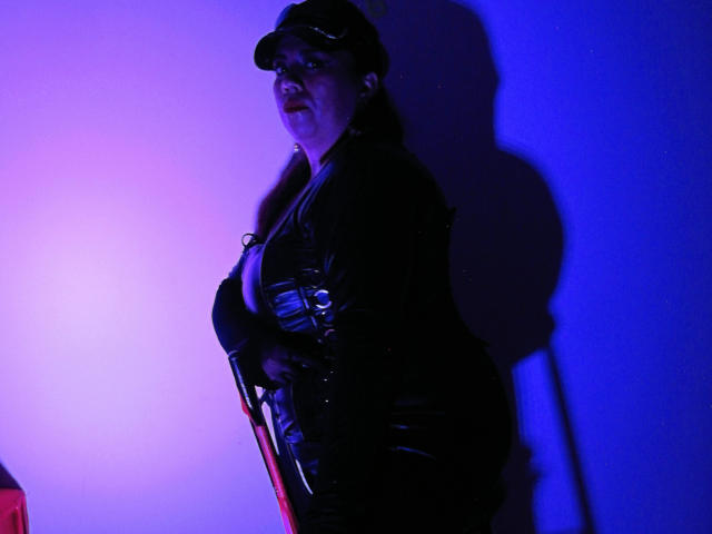 LexyDom con un outfit in pelle scura, un cappello e delle cinghie, in piedi in una luce viola-blu, che mostra il sedere, tenendo un bastone rosso, con un'ombra sul muro..