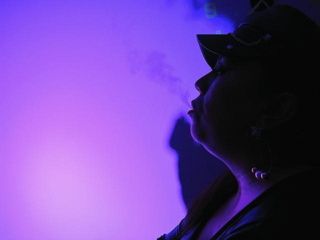 LexyDom with a side profile, dark silhouette, purple-blue light, smoking, wearing a cap, earrings, and a jacket, shadow of her head on the right.