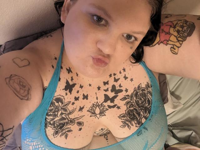 MilaSexy69Hot with blue mesh top, showing large tits, tattoos of butterflies, roses, and a heart on her chest and arms, pouting with dark hair, lying on bed.