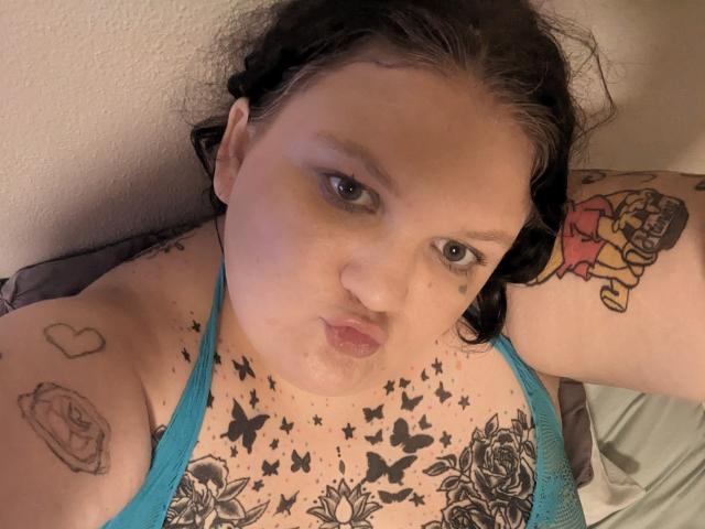 MilaSexy69Hot with black tattoos on her chest, arms, and neck, wearing a blue tank top, lying on a bed, looking up with pouty lips, dark hair.
