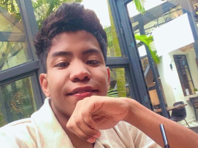 ZiziNine has a cute face, brown curly hair, and is smiling with their chin resting on their hand. They're wearing a striped shirt. The image is a selfie.